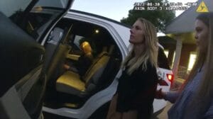 Two women stand next to a police vehicle at dusk; one is being guided by another woman, while a third person sits inside the back seat. The scene appears to be recorded by a body camera.