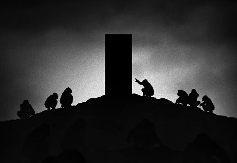 Silhouetted figures resembling apes gather around a tall, rectangular monolith on a hilltop, dramatically lit from behind, creating a mysterious and intense scene from the film.