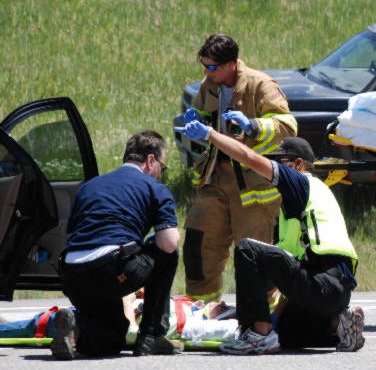 Randy working a crash on the highway. He is kneeling over a patient who has been extricated from a crashed car. Another medic is gesturing while a firefighter looks on.