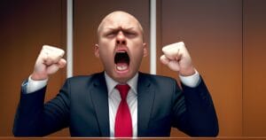 A bald man in a suit and red tie sits at a desk, clenching his fists and yelling with his mouth wide open, appearing very angry or frustrated. The background is plain and brown, like a courtroom.