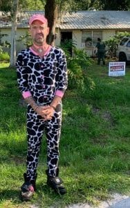 A man wearing a cow-print onesie, pink cap, and black sneakers stands on grass in front of a house with a 'This drug house is closed' sign in the yard. A police officer is near the house in the background.