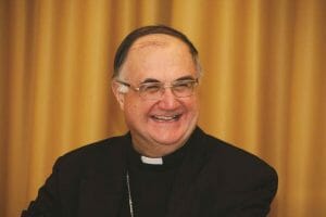 A chuckling priest wearing glasses, a black clerical suit, and a white clerical collar in front of a golden yellow curtain.