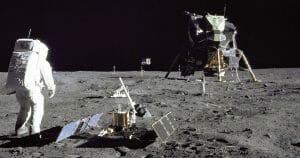 Buzz Aldrin stands on the Moon’s surface near scientific equipment, with the Apollo 11 lunar module visible in the background, under a black sky.