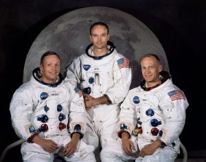 The three Apollo 11 astronauts in their white NASA spacesuits pose for their official portrait in front of a large image of the moon, smiling and seated together.