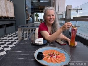 A woman with long gray hair sits at a rooftop restaurant table, smiling and holding a tall, colorful drink. In front of her is a plate of thinly sliced salmon sashimi with garnish and sauce. The backdrop includes boats scattered across the sea.