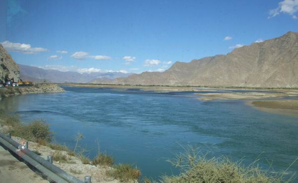 A wide, calm river flows between dry, rocky mountains under a clear blue sky. Sparse vegetation lines the riverbank, and a metal guardrail runs along the roadside in the foreground.