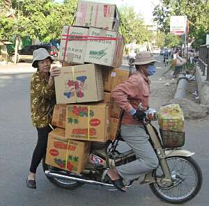 Loaded with boxes — plus one wife. Two people ride a small motorbike overloaded with large cardboard boxes, some stacked high and tied to the bike. The passenger helps steady the boxes, and both wear hats. They are on a street with trees and other vehicles.