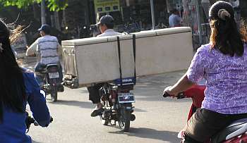 Carrying a refrigerator Four people ride motorbikes on a street; one carries a large refrigerator strapped behind, while the others ride normally. Sunlight casts shadows on the road.