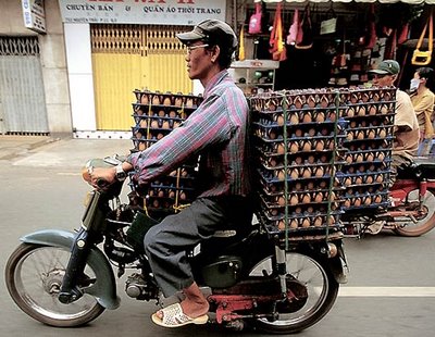 A large load of eggs. A man rides a motorcycle on a city street, carrying large racks filled with eggs stacked on both sides and behind him. He wears a checkered shirt, cap, sunglasses, and sandals.