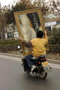 A large painting. A person riding on the back of a motorcycle on a street while awkwardly carrying a large, framed painting. The painting is tilted sideways and obstructs the rider’s view. The driver is sitting in front of the large painting. Trees and buildings are visible in the background.