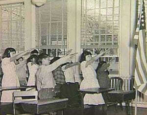 A group of schoolchildren in a classroom stand with their right arms extended toward an American flag at the front of the room, reciting the Pledge of Allegiance. The image is in black and white.