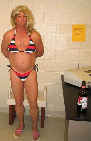 A police photo of a man wearing a blond wig and a striped bikini and flip-flops stands indoors next to a table with a beer bottle and office equipment, facing the camera with hands behind his back. He has a bit of a beer belly, and is looking a bit sullen.