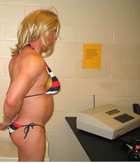 A police photo of a man wearing a blond wig and a striped bikini stands indoors next to a table with a beer bottle and office equipment, facing the camera with hands behind his back. in front of a machine on a counter, in a room with beige brick walls and a yellow sign posted on the wall. His bikini top appears to be stuffed with one orange on each side. We can see he is handcuffed behind his back.