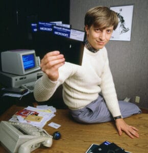 Bill Gates in a white sweater sits on a desk holding up three floppy disks labeled Microsoft. A vintage computer, papers, and office supplies are visible on the desk in a 1980s office setting.