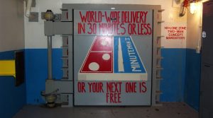 A large metal vault door painted with WORLD-WIDE DELIVERY IN 30 MINUTES OR LESS OR YOUR NEXT ONE IS FREE and MINUTEMAN II, referencing a missile system. A sign on the wall reads NO-LONE ZONE TWO-MAN CONCEPT MANDATORY.