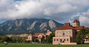 The University of Colorado campus in Boulder with stone buildings and red roofs sits in front of green fields and trees, with the dramatic Rocky Mountains in the background and low clouds above.
