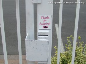 A white metal gate features a sign with pink text reading Thanks for Cumming! above a graphic of humping bunnies. The gate is next to a small plant and an asphalt surface.