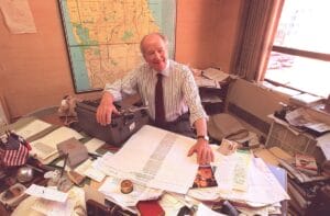 An older man in a striped shirt smiles at a cluttered desk with papers, books, and a typewriter. A sunlit map of San Francisco is on the wall behind him.