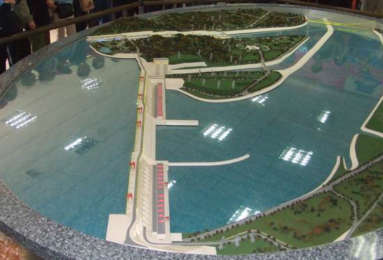 A detailed scale model of a hydroelectric dam with surrounding green landscape, water reservoir, and small buildings, displayed on a round table with people observing in the background.