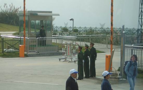 Three uniformed guards stand near a security gate, with another guard in a booth nearby. Three civilians, one wearing a blue jacket and two others wearing white caps, walk past the area. Fences and greenery are visible in the background.