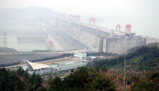 A large hydroelectric dam with several red cranes spans a wide river, surrounded by mist. In the foreground, there are green hills and buildings with trees and vegetation. The air is heavily polluted.