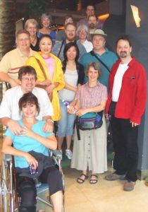 A group of fifteen adults, including a woman in a wheelchair, pose together indoors, smiling at the camera. Most are casually dressed, standing on steps in a well-lit area.