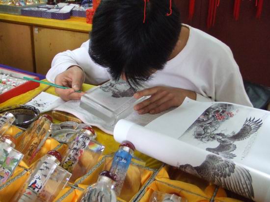 A person with short black hair uses a magnifying glass and a thin brush to paint intricate designs inside a glass bottle, referencing an open book with an eagle illustration, surrounded by decorated bottles.