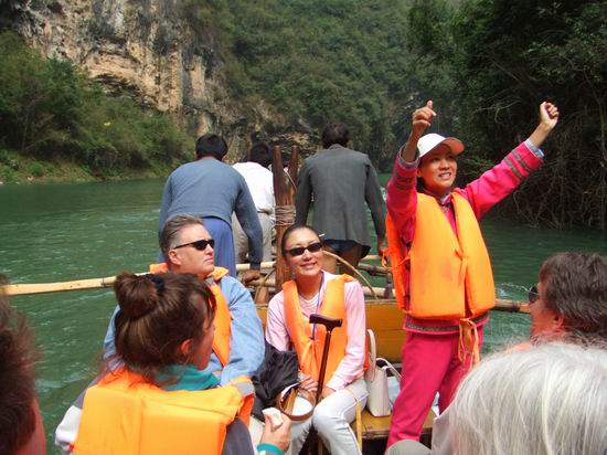 A group of people wearing orange life jackets ride a wooden boat along a river surrounded by rocky cliffs and greenery. One person stands at the front, smiling and raising both arms in excitement. At the front of the boat, three men look intently forward as there are the ones who jump out and pull the boat through narrow channels.