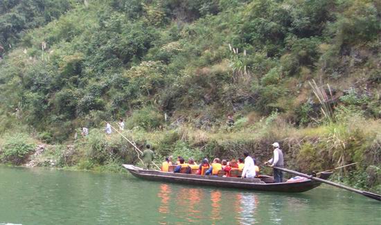 A group of people wearing life jackets sit in a long wooden boat on a green river, surrounded by lush, green hills and dense vegetation. A person stands at the back, steering the boat with a pole, while several men on a path on shore pull the boat along with a rope.