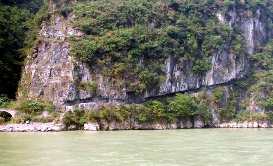 A steep, rocky cliff covered in green vegetation rises beside a wide, calm river. A small path is carved into the base of the cliff along the riverbank.