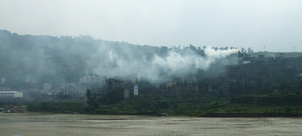 Smoke rises from industrial buildings near a river, with green hills and scattered structures in the background under an overcast sky. The scene suggests air pollution in a developing urban area.