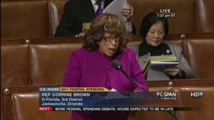 Rep. Corrine Brown, wearing a purple jacket, speaks at a lectern in the U.S. House chamber. Another woman sits behind her reading papers. The screen displays information about federal spending and C-SPAN coverage.