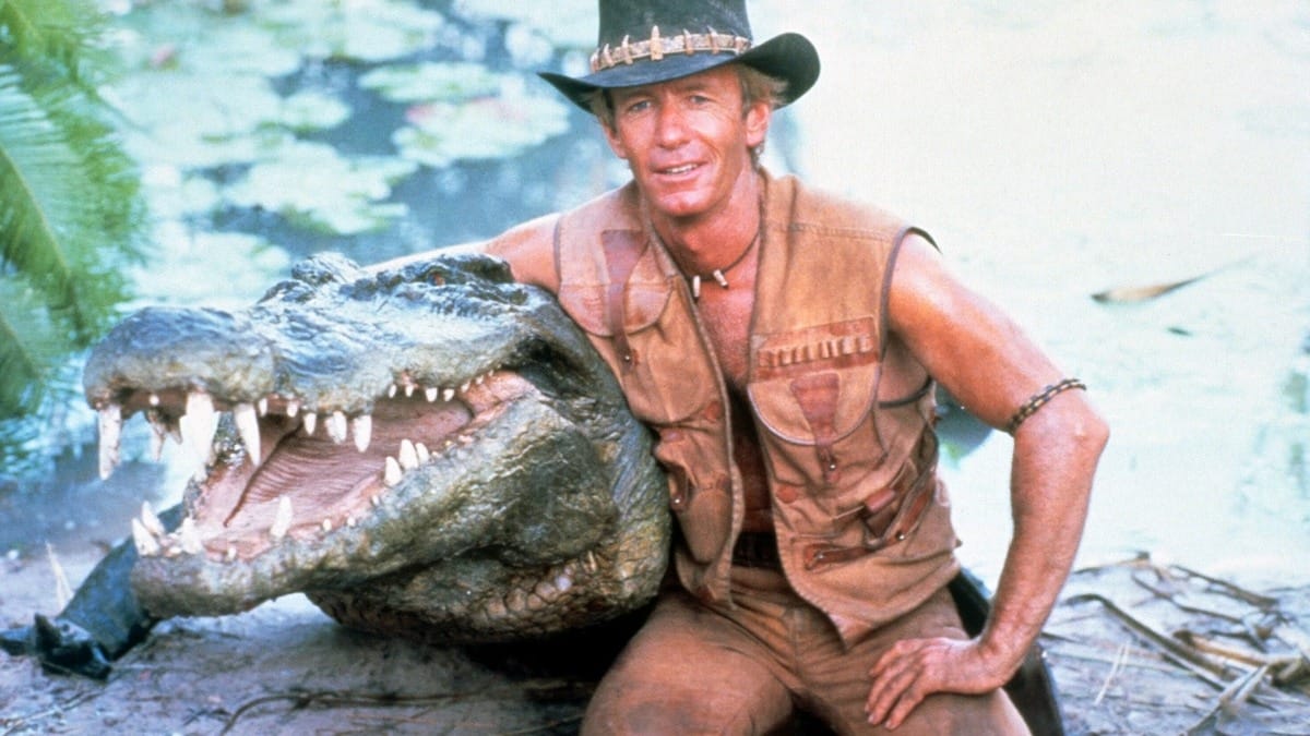 Actor Paul Hogan, wearing a brown leather vest and hat, kneels beside a large crocodile with its mouth open, set outdoors near water.