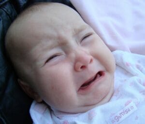 A baby boy with light skin and light brown hair is crying loudly with its mouth wide open. Tears are on his cheeks, and his eyes are squeezed shut. The background is plain white.