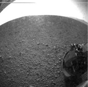 Black and white image depicts a rocky, dusty surface that's abnormally curved due to lens distortion, reflecting it as a test image. A visible mechanical arm indicates the photo was captured by a spacecraft exploring Mars.