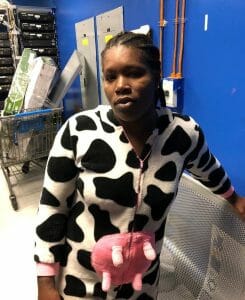 A different woman standing indoors, wearing a black and white cow-print onesie with a pink udder design, leaning against a metal bench. There are shelves, equipment, and a blue wall in the background.