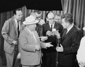 A group of reporters standing in front of a curtain on a stage clustered around Khrushchev and Richard Nixon as they conduct the 'Kitchen Debate' on July 24, 1959. Newsman John Charles Daly is most prominent among the reporters.