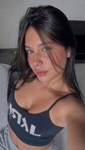 A young woman with long dark hair wearing gray sweatpants and a black sports workout bra. She looks straight at the camera in a selfie pose.