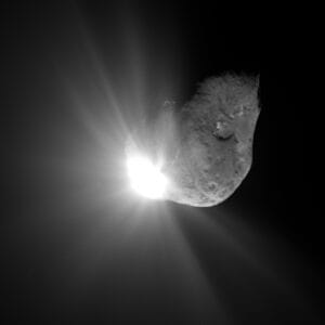 A bright burst of light illuminates part of a lumpy, irregularly shaped comet in space, with rays and shadows radiating outward against a dark background.