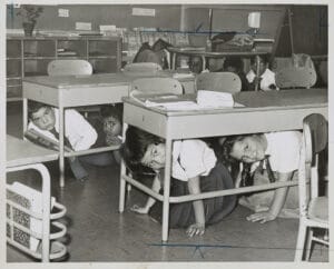 Schoolchildren crouch under their classroom desks during a 'duck and cover' drill, smiling at the camera. Books and supplies are scattered on the desks.