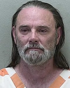 A mug shot of a middle-aged man with long brown hair, a full gray beard, and mustache is wearing an orange and white striped prison uniform against a plain light-colored wall. He has refused to look at the camera.