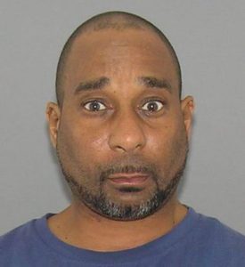 A mug shot of a man with short hair and a goatee wearing a blue shirt stands in front of a plain gray background, looking directly at the camera with a surprised expression.