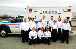 A group of thirteen people in dress white uniform shirts stand and kneel in front of an OCEMS ambulance parked outdoors, posing for a group photo.