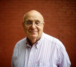 Dave Farber with glasses and a receding hairline smiles wryly, wearing a striped button-down shirt and standing in front of a red brick wall.