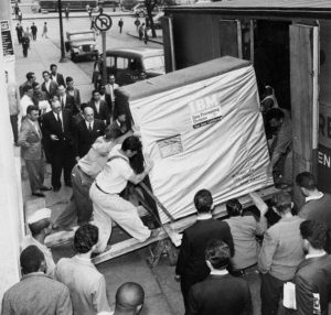 A group of men push a large, covered box labeled IBM Data Processing System up a ramp into a building as a crowd watches on a city sidewalk in the 1950s.