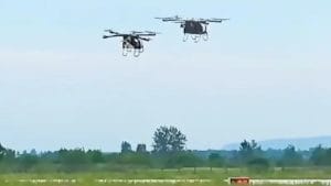 Two black drone-like flying vehicles hover above a grassy field with trees and distant hills in the background under a clear sky.
