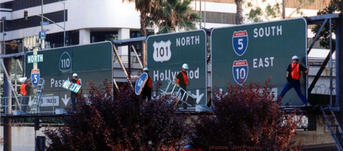 Construction workers in safety vests and helmets work on large freeway signs for Interstate 5, 110, and 101, surrounded by trees with a city building in the background.