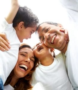 A happy family of four, two adults and two children, wearing white shirts and smiling joyfully as they embrace and laugh together. The photo is taken from below, looking up at their faces.