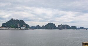 Cloudy sky over calm water with green, rocky islands in the distance. Along the shore at the base of a large, forested hill, there are buildings and boats.