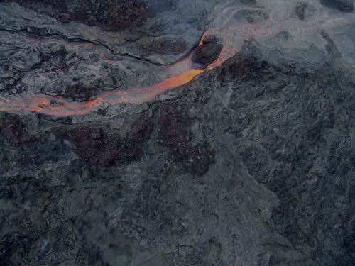 Aerial view of dark, rocky terrain with a glowing orange stream of molten lava flowing through cracks on the surface.